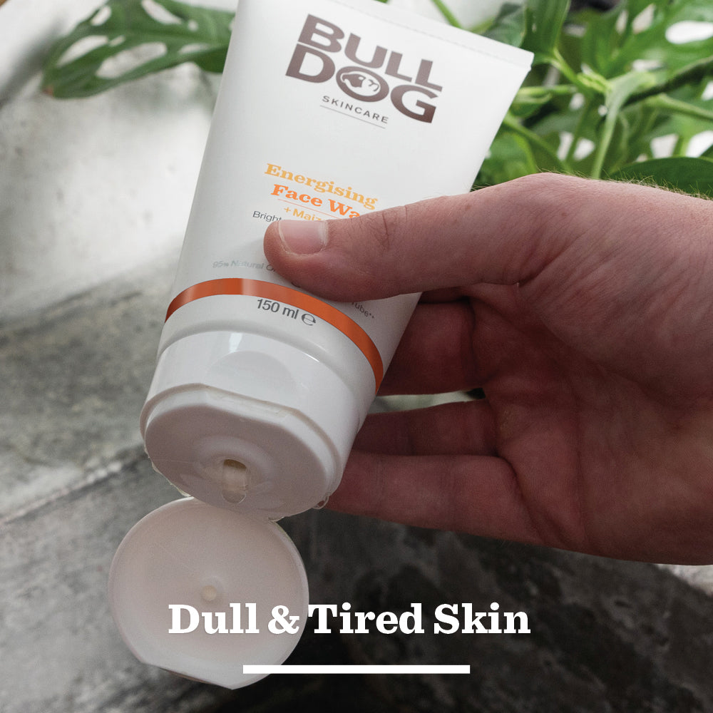A hand squeezes a tube of Energising Face Wash for men with an open cap, set against green plants. Text reads, “Dull & Tired Skin.” Crafted with natural origin ingredients to revitalise tired skin.