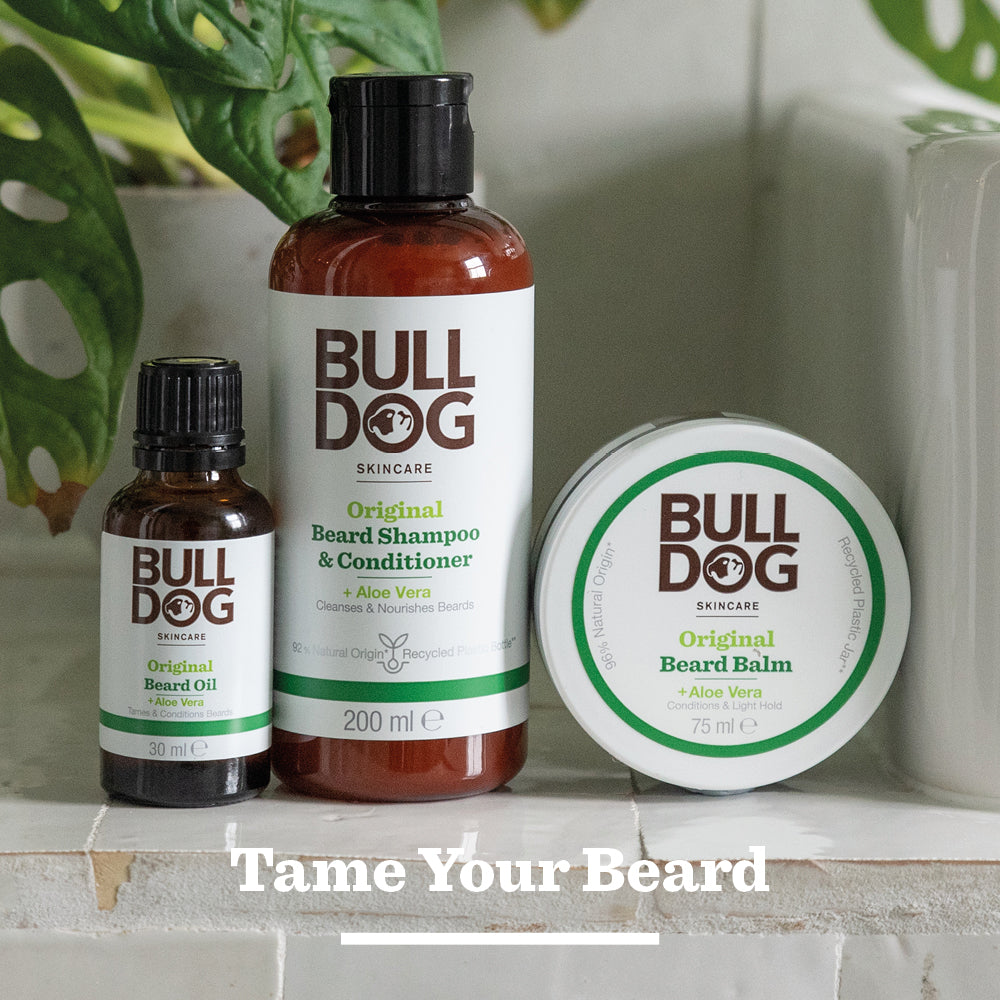 Bulldog Skincare’s Original Beard Balm, made with natural origin ingredients, sits on a bathroom counter with green leaves in the background. Text on image: Tame Your Beard.