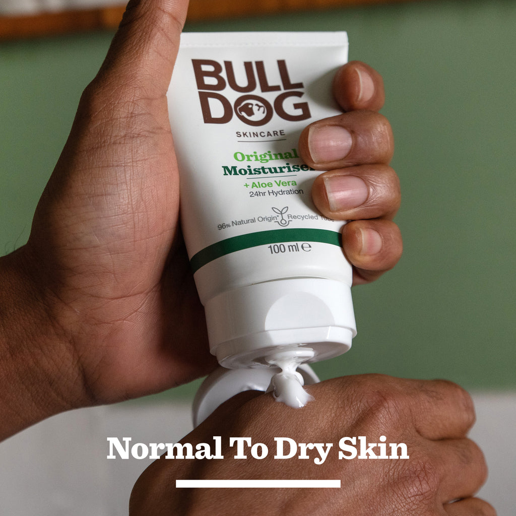 A hand squeezes a tube from the Original Skincare Trio with Aloe Vera. The label states 24hr hydration, suitable for normal to dry skin. Perfect Bulldog gift for men; the background features a blurred green wall.